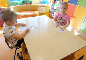 Dwoje dzieci siedzi przy stole i składa z części obrazek łąki.
